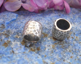 May include: Two small, silver-colored metal beads with a hammered texture. The beads are cylindrical with a hole through the center. The beads are displayed on a blue surface with purple flowers in the background.