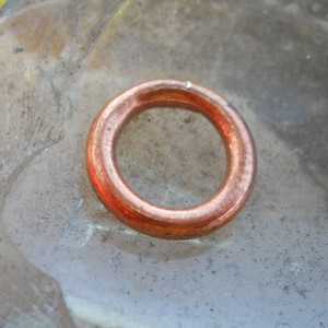 May include: A copper-coloured, circular washer with a hole in the centre. The washer is made of a shiny, metallic material and is resting on a mottled, grey surface. The washer is a component used in plumbing or mechanical applications.