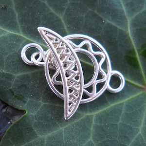 May include: Silver-toned metal clasp with a sun and moon design. The clasp is intricate and detailed, with a circular sun design and a crescent moon design.