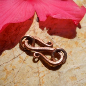 May include: A copper-colored metal clasp with a unique double-loop design. The clasp is lying on a light brown surface.