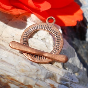 May include: A copper-colored metal toggle clasp with a circular design and a textured surface. The clasp has a loop on the top for attaching to a necklace or bracelet.