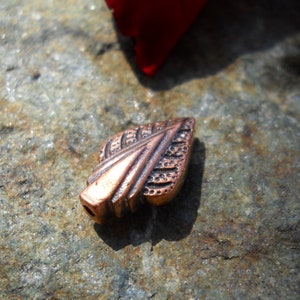 May include: A copper leaf-shaped bead with a detailed design. The bead has a textured surface and a pointed tip.