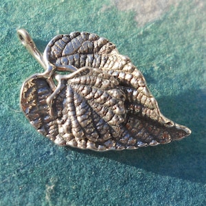 May include: A silver leaf pendant with intricate veining and a textured finish. The pendant shows two overlapping leaves and a small loop for a chain. The design is inspired by nature and suitable for jewellery.