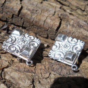 May include: Two silver metal square-shaped clasps with intricate swirl designs and a raised square in the center. The clasps have a loop on the bottom for attaching to a necklace or bracelet.