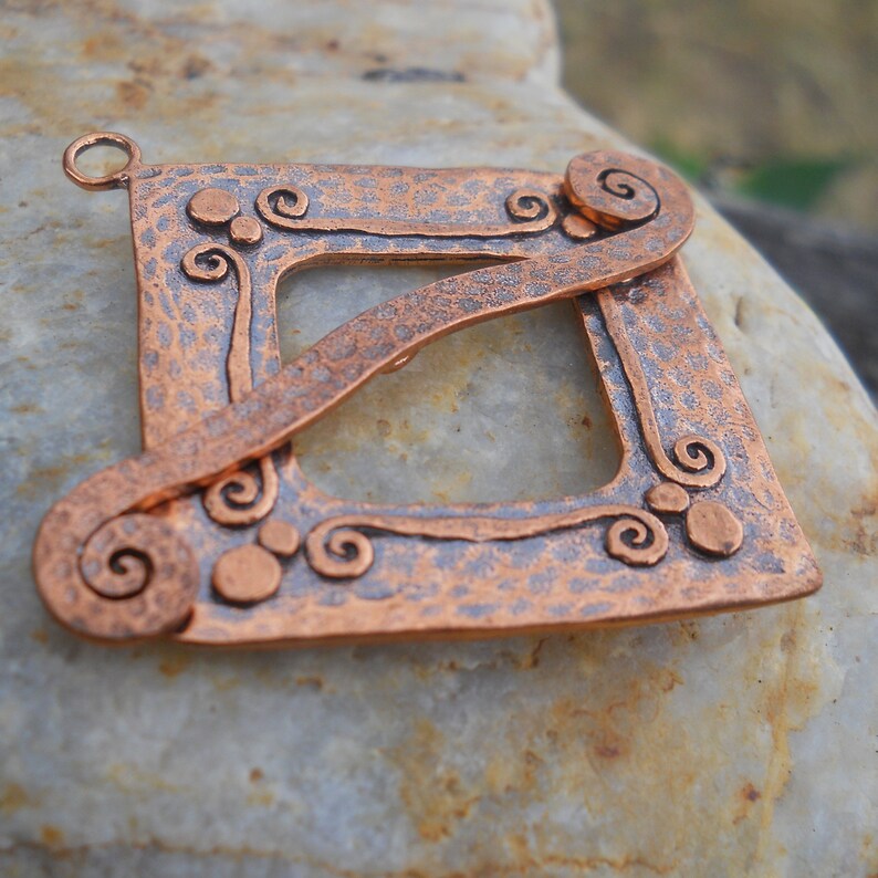 Op de afbeelding: Een gehamerde koperen hanger met een vierkant frame en een zwierig ontwerp. De hanger heeft een getextureerd oppervlak en een lus aan de bovenkant om op te hangen.