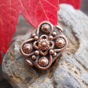 Puede incluir: Una cuenta de metal de color cobre con un diseño floral decorativo. La cuenta tiene un centro elevado con un patrón circular y está rodeada de círculos más pequeños.