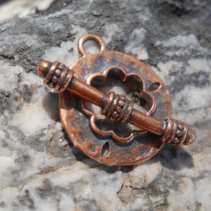 May include: A copper-toned metal clasp with a decorative flower design. The clasp has a round shape and a bar that goes through the center. The clasp is designed for jewelry making.