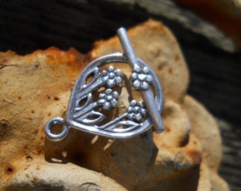 May include: Silver-toned metal clasp with a floral design. The clasp is shaped like a teardrop with a bar that slides through the loop. The floral design features delicate leaves and flowers.
