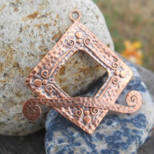 Op de afbeelding: Een koperen hanger met een vierkant frame en een gehammerde textuur. Het frame heeft een uitsparing in het midden en is versierd met swirls en stippen. De hanger heeft een lus aan de bovenkant om een ketting aan te bevestigen.