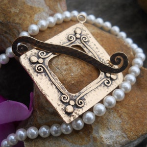 May include: A gold-toned pendant with a square frame and intricate scrollwork design. The pendant is attached to a strand of white pearls.