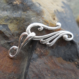 May include: A silver-colored jewelry clasp with an ornate, flowing design. The clasp features a hook and eye closure with curved, scroll-like details. The metal has a polished finish, reflecting light.