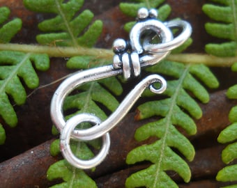 May include: Silver-toned metal hook clasp with a decorative design. The hook is closed and has a rounded end. The clasp is photographed against a green fern background.