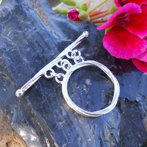 Puede incluir: Cierre de palanca de plata con un bucle redondo de alambre retorcido. El cierre está sobre un fondo gris oscuro.