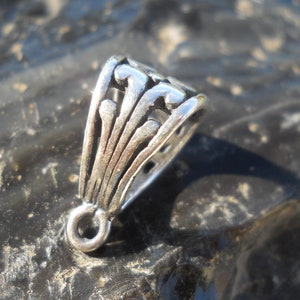 May include: Silver pendant bail with a decorative, ornate design. The bail has a loop at the bottom for attaching a pendant.