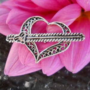 Puede incluir: Una pinza para el pelo de plata filigranada en forma de corazón con un diseño decorativo trenzado en forma de cuerda. La pinza está apoyada sobre un pétalo de flor rosa.