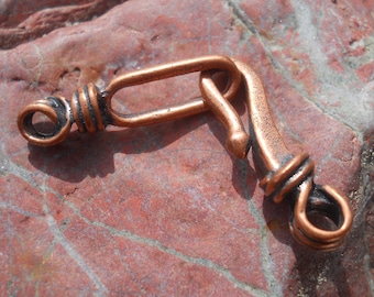 May include: A copper-colored clasp with an oval loop and a hook closure. The clasp has a twisted wire design on both ends and is set against a reddish-brown stone surface. The metal has a slightly aged appearance.