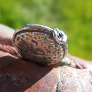 May include: A silver charm in the shape of a small, round, textured disc with a raised, curved, textured detail on the side.