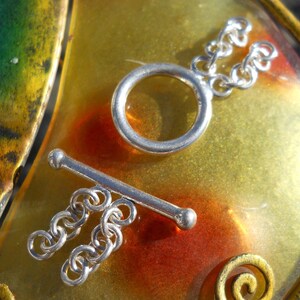 May include: Silver toggle clasp with a round ring and a bar. The clasp is on a textured, yellow, orange, and green surface.