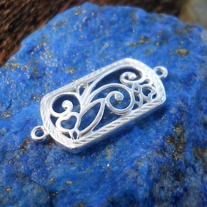 May include: A silver filigree connector with an ornate, openwork design. The rectangular piece features swirling patterns and loops within a textured border. Small loops are on each end for attachment. The connector is displayed on a blue, textured surface.