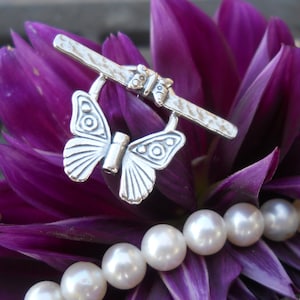 Puede incluir: Cierre de mariposa de plata con una barra texturizada. El cierre está sobre una flor morada con perlas blancas.