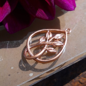 May include: A rose gold pendant charm featuring a stylised tree branch with leaves. The charm is in the shape of a teardrop and has a loop at the top for attaching to a necklace or bracelet.