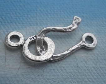 May include: Silver clasp with a textured, hammered finish. The clasp is in the shape of a loop with a smaller loop on one side and a hook on the other. The clasp is marked "925" indicating it is made of sterling silver.