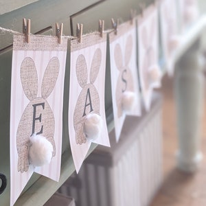Può includere: Uno striscione con la parola "PASQUA" scritta con bandierine a forma di coniglio. Ogni bandierina ha un coniglio marrone con una coda di cotone bianco e uno sfondo a righe rosa e bianco.