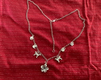 May include: A silver chain necklace with multiple charms, including a flower, a heart, and a dog. The necklace is on a red fabric background.