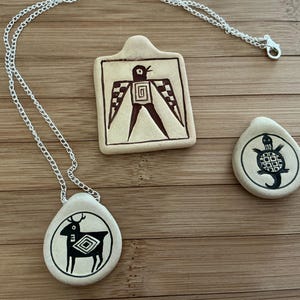 May include: Three ceramic pendants with silver chains. One pendant is a rectangle with a bird design, another is a teardrop shape with a turtle, and the third is an oval with a deer. All designs are black on a cream background.