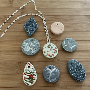 May include: A collection of handmade ceramic pendants in various shapes and colors, including a diamond, circles, and teardrop. The pendants feature embossed designs and are strung on a silver chain. Some pendants have red and green accents.