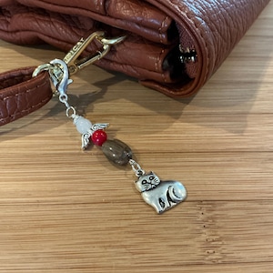 Puede incluir: Un bolso de cuero marrón con un dije de plata adjunto. El dije presenta un pequeño ángel con alas blancas, cuentas rojas y un gato plateado.