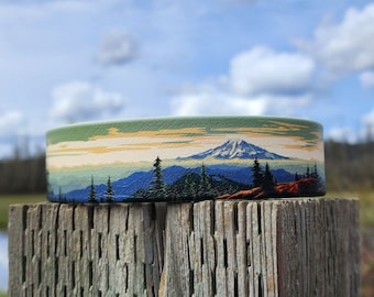 May include: A green, blue, and white mountain landscape printed on a fabric wristband. The wristband is sitting on a weathered wooden surface.