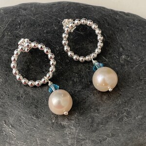 May include: A pair of silver hoop earrings with a pearl and blue crystal dangle. The hoops are made of small silver beads.