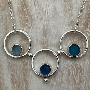 May include: A silver necklace with three circles, each containing a different shade of blue sea glass.