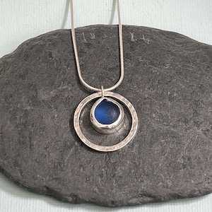 May include: A silver necklace featuring a hammered-texture pendant with a teardrop-shaped, deep blue glass stone. The pendant is encircled by a larger hammered silver ring. The necklace is displayed on a dark gray stone surface.