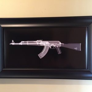 May include: Framed print featuring a detailed, x-ray style illustration of an AK-47 assault rifle. The image is rendered in shades of white and gray against a stark black background. The print is encased in a black rectangular frame.