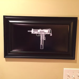 May include: Framed artwork featuring a white, x-ray style image of a submachine gun against a black background. The artwork is encased in a black rectangular frame, creating a striking contrast. The piece is mounted on a light yellow wall.