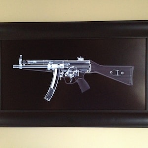 May include: Framed artwork featuring an x-ray style image of a submachine gun. The gun is depicted in white against a black background, showcasing its internal components. The artwork is encased in a black frame.