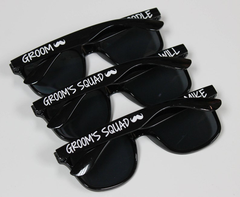 May include: Three black sunglasses with white text. The top pair says "Groom" with a mustache symbol. The middle and bottom pairs say "Groom's Squad" with a mustache symbol.