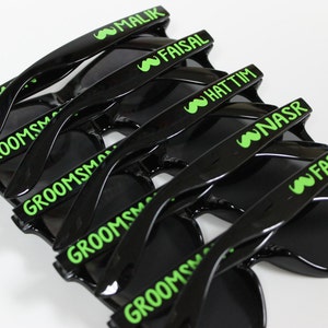 May include: A stack of black sunglasses with green text. The text on the sunglasses reads "Best Man", "Groomsman", "Malik", "Faisal", "Hattim", "Nasr", and "Farris".