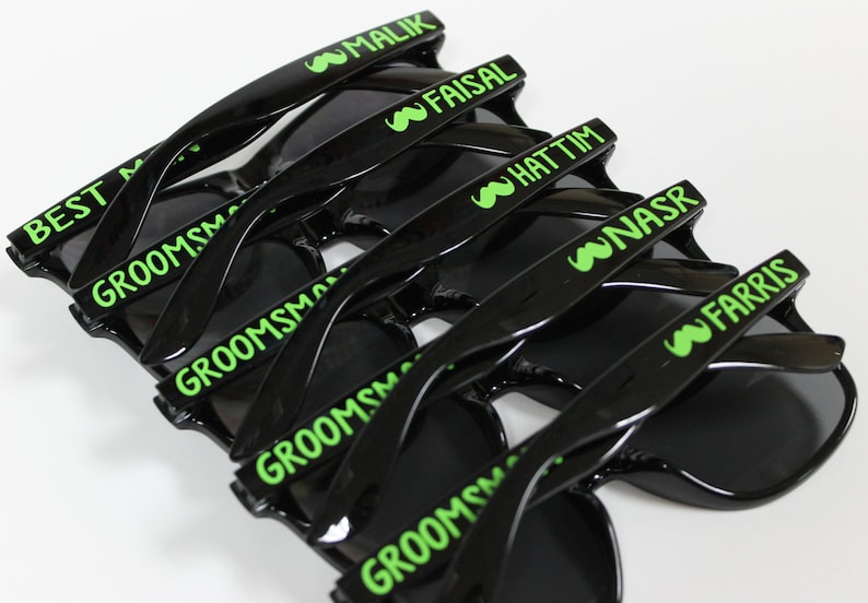 May include: A stack of black sunglasses with green text. The text on the sunglasses reads "Best Man", "Groomsman", "Malik", "Faisal", "Hattim", "Nasr", and "Farris".