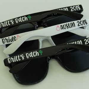 May include: Three pairs of sunglasses with black frames and dark lenses. The top pair has the text "Britt's Bach" with a green cactus icon. The middle pair has the text "Bride" with a pink ring icon. The bottom pair has the text "Austin 2018" with a pink ring icon.