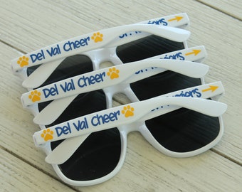 May include: Three white sunglasses with blue and yellow text that reads "Del Val Cheer" on the arms. The sunglasses have black lenses.