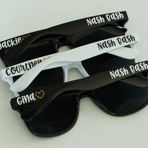 May include: Three pairs of sunglasses with different names printed on them. The top pair is black with white text that says "Nash Bash". The middle pair is white with black text that says "Nash Bash". The bottom pair is black with white text that says "Gina" with a gold heart symbol.
