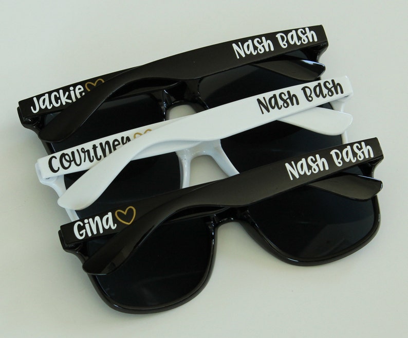 May include: Three pairs of sunglasses with different names printed on them. The top pair is black with white text that says "Nash Bash". The middle pair is white with black text that says "Nash Bash". The bottom pair is black with white text that says "Gina" with a gold heart symbol.