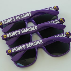 May include: Three pairs of purple sunglasses with black lenses. The sunglasses have a white inscription that says "Bride's Beaches" and a yellow sun icon. The arms of the sunglasses have a yellow inscription that says "Sarah", "Jessie", and "Lissa" with a yellow champagne glass icon.