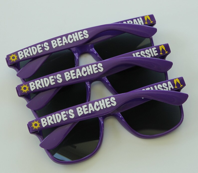 May include: Three pairs of purple sunglasses with black lenses. The sunglasses have a white inscription that says "Bride's Beaches" and a yellow sun icon. The arms of the sunglasses have a yellow inscription that says "Sarah", "Jessie", and "Lissa" with a yellow champagne glass icon.