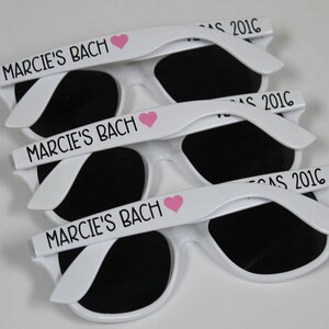 May include: Three white plastic sunglasses with black lenses. Each pair has the text "Marcie's Bach" and a pink heart printed on the frame.