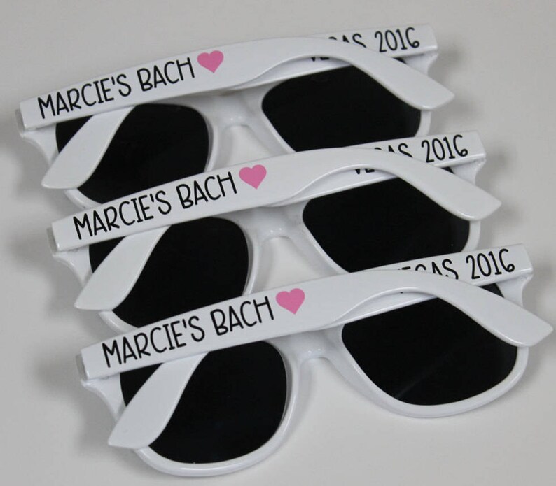 May include: Three white plastic sunglasses with black lenses. Each pair has the text "Marcie's Bach" and a pink heart printed on the frame.