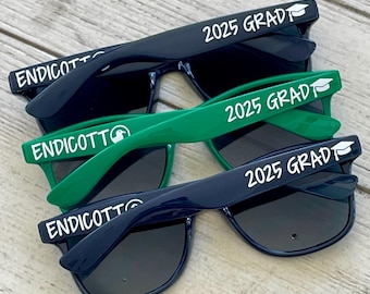 May include: Three pairs of sunglasses with black frames and dark lenses. The sunglasses have white text on the frames that says "Endicott" and "2025 Grad" with a graduation cap icon. The sunglasses are stacked on top of each other on a wooden surface.